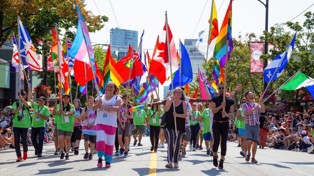 People marching in a Pride parade in downtown Vancouver and carrying the pride flag, trans flag, Canada flag, and flags of Canadian provinces and territories.