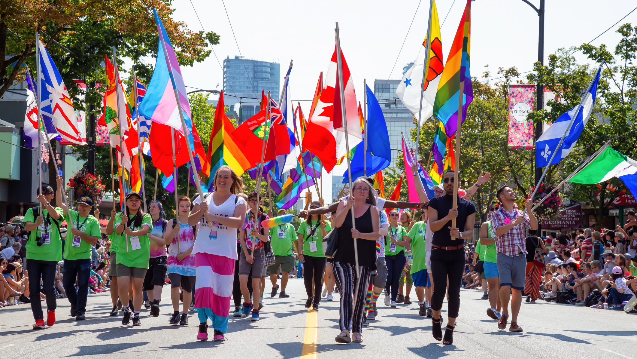 Dans le centre-ville de Vancouver, des participants au Défilé de la fierté brandissent des drapeaux arc-en-ciel, des drapeaux de la fierté transgenre et des drapeaux du Canada et des provinces et territoires canadiens.