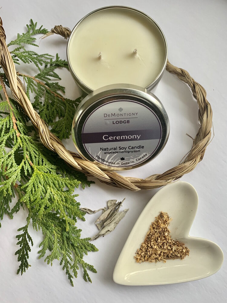 Une boîte argentée ouverte contenant une bougie de soya de couleur crème placée à côté de branches de cèdre, de la ficelle tissée et un plat de couleur crème en forme de cœur contenant des herbes.