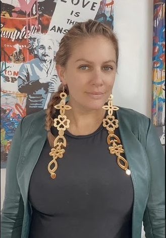 Blonde woman wearing a dark grey top, pale green leather jacket and very long, ornate gold earrings looking straight into the camera with colourful artwork behind her.