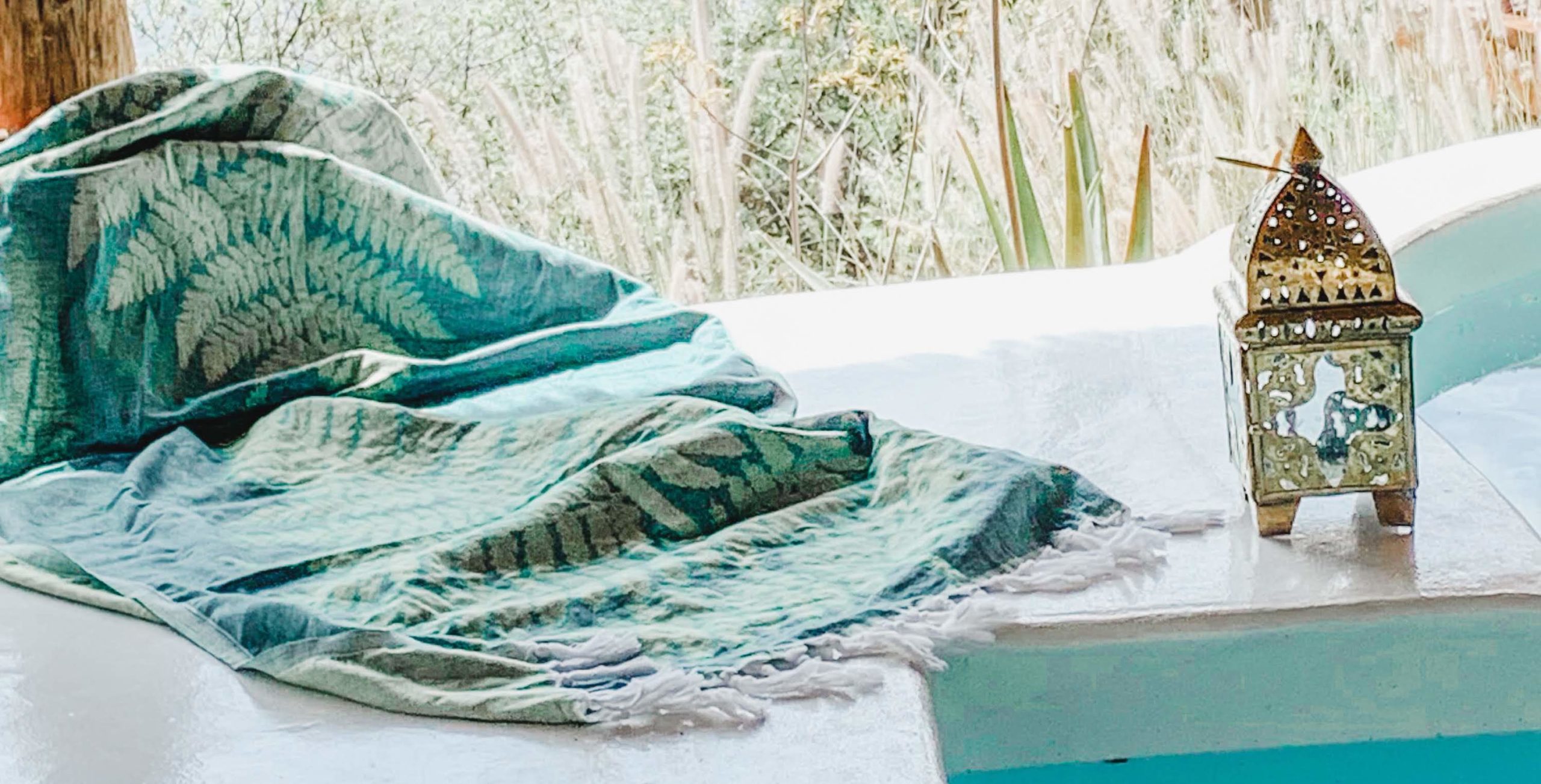 A light turquoise, Turkish woven blanket with cream coloured fern print and long tassel fringe laid upon the edge of a bath with a Turkish bronze lantern.