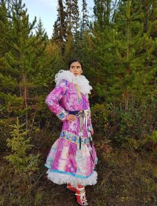 Indigenous woman wearing a long, bright, multi-coloured patchwork parka and red mukluks looking into the distance in the forest.