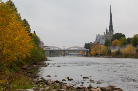 A dark grey church on the horizon on the right, arched bridge in the middle with the river flowing underneath, surrounded by yellow and green foliage on the embankments.