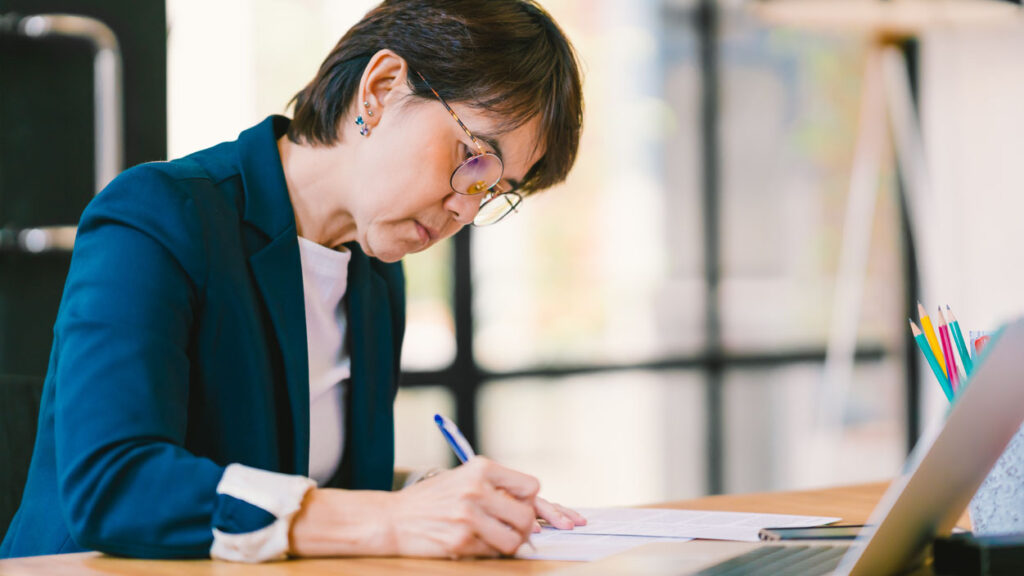 Portrait d’une femme en blazer bleu assise à un bureau, en train d’écrire sur une feuille de papier, avec un ordinateur portatif au premier plan