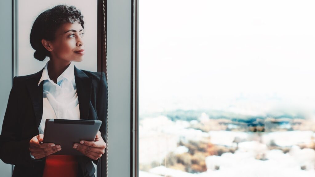Femme tenant une tablette, adossée à une grande fenêtre et contemplant un paysage urbain lumineux