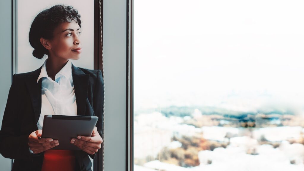 A woman holding a tablet and leaning against a large window, looking out over a bright cityscape