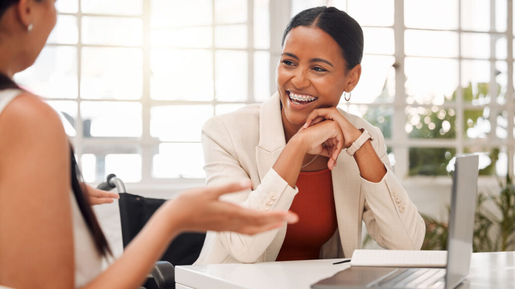 Two women in business attire and talking by a desk and computer