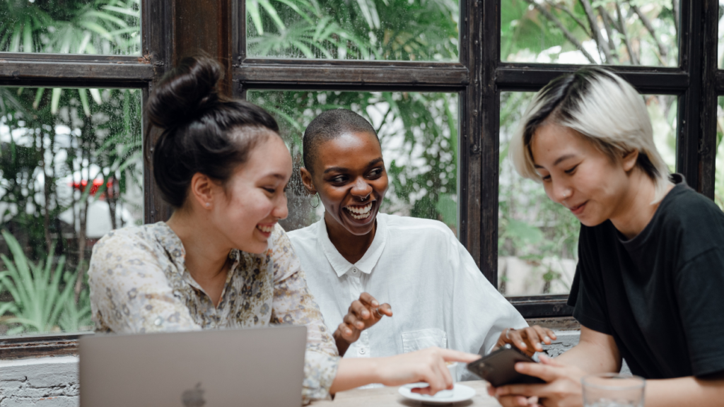 Three diverse women at a table smiling with two of them looking at a phone.