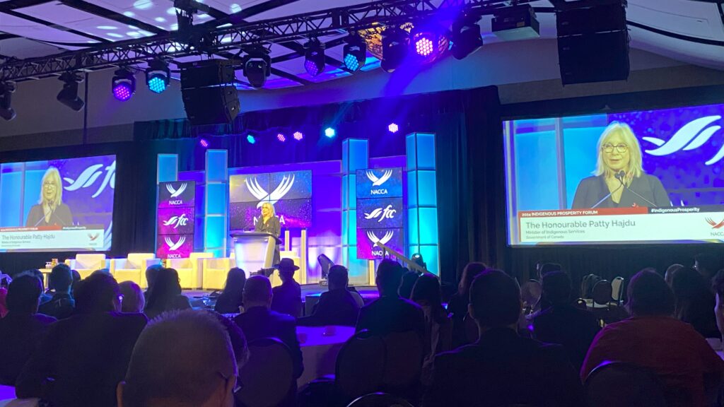 A woman speaking at a podium on a stage with her speech broadcast on two large screens and the logos of NACCA on the screens behind her.