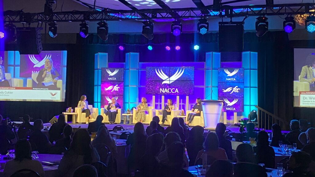 Five women sitting on stage during a panel discussion with logos of NACCA in the background.