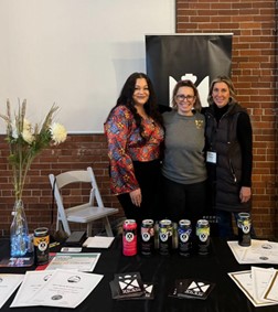 Three women pose and smile in front of a table with cans of beer.