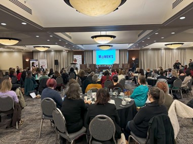 A wide-angle view of the conference room with attendees sitting at round tables, facing a stage at the front.