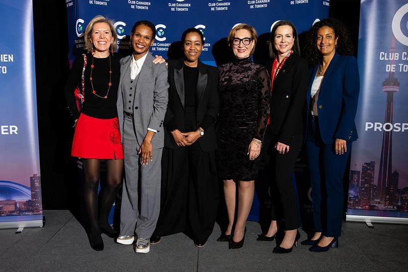 Six diverse women posing together at a professional event, standing in front of a blue backdrop with "Club Canadien de Toronto" branding. They are dressed in business and formal attire, smiling and standing closely together.