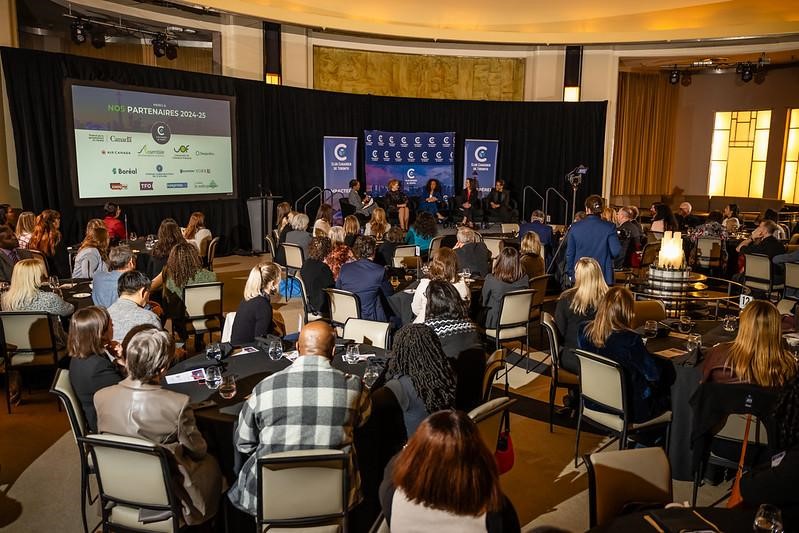 A large audience attending a panel discussion at the Club Canadien de Toronto event. The attendees are seated at round tables, listening to the speakers on stage. A large screen displays the event’s partners.